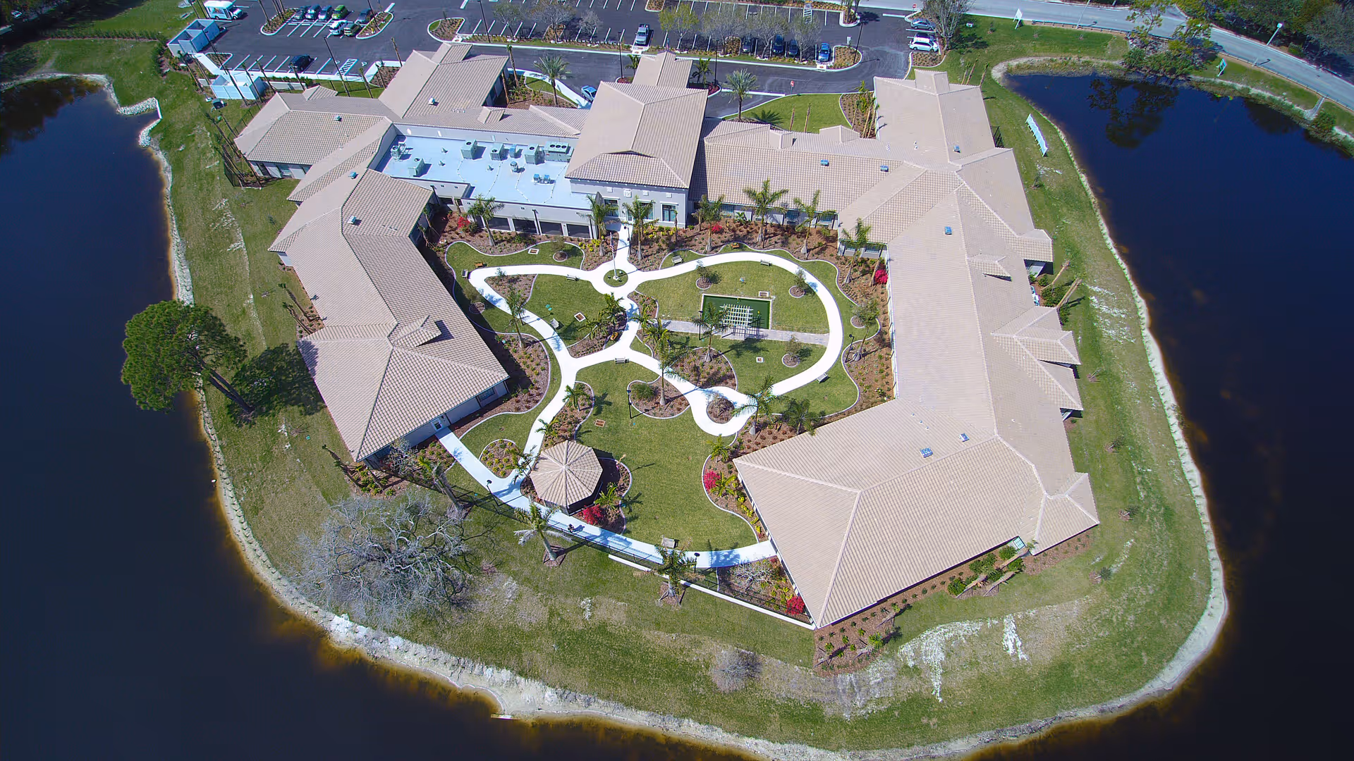Aerial view of a senior living facility building surrounded by a water body, featuring a landscaped garden with winding white pathways, palm trees, and a gazebo in the center courtyard. The building has a beige roof and is adjacent to a parking lot.