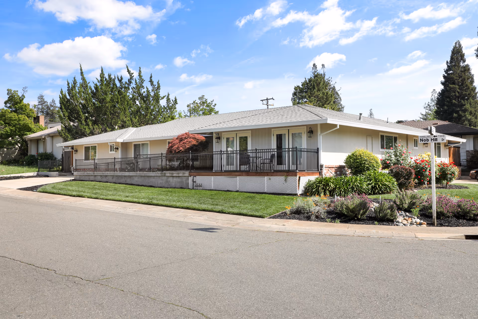 Exterior view of a single-story residential building with a light-colored facade and a gray roof. The building has a raised porch with black railings and outdoor chairs. There is a well-maintained lawn and landscaped garden beds with various shrubs and flowers. A street sign at the corner reads 'Nob Hill Dr'. The sky is blue with scattered clouds.