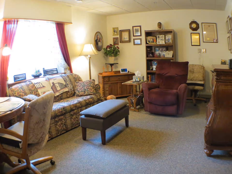 A cozy living room with a floral patterned sofa, a burgundy recliner chair, a gray ottoman, and a wooden side table with tissues and small items. The room has framed pictures and certificates on the walls, a wooden bookshelf filled with books and photos, a floor lamp, and a window with red curtains letting in natural light.