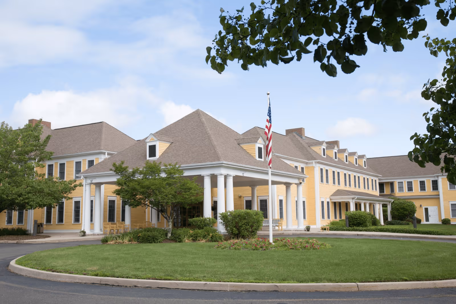 Exterior view of a large yellow senior living facility building with white columns at the entrance, an American flag on a flagpole in front, surrounded by green trees and a circular driveway.