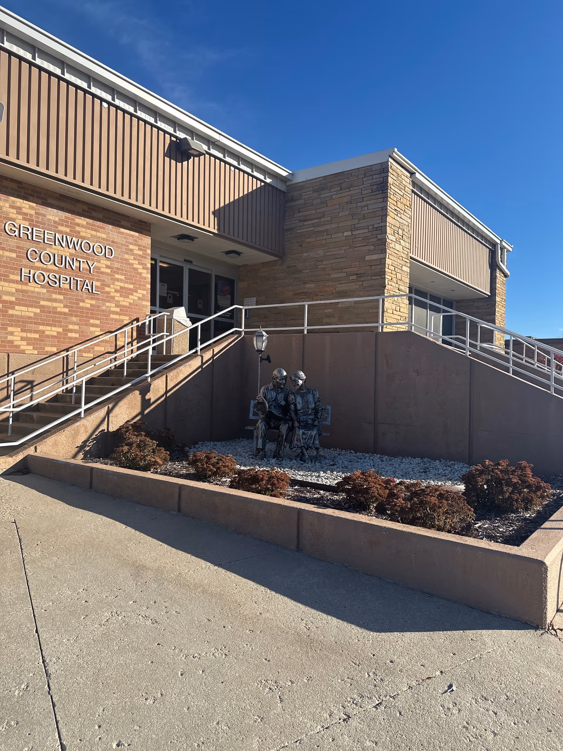 Front exterior of Greenwood County Hospital with ramp, brick facade, and seated sculptures.