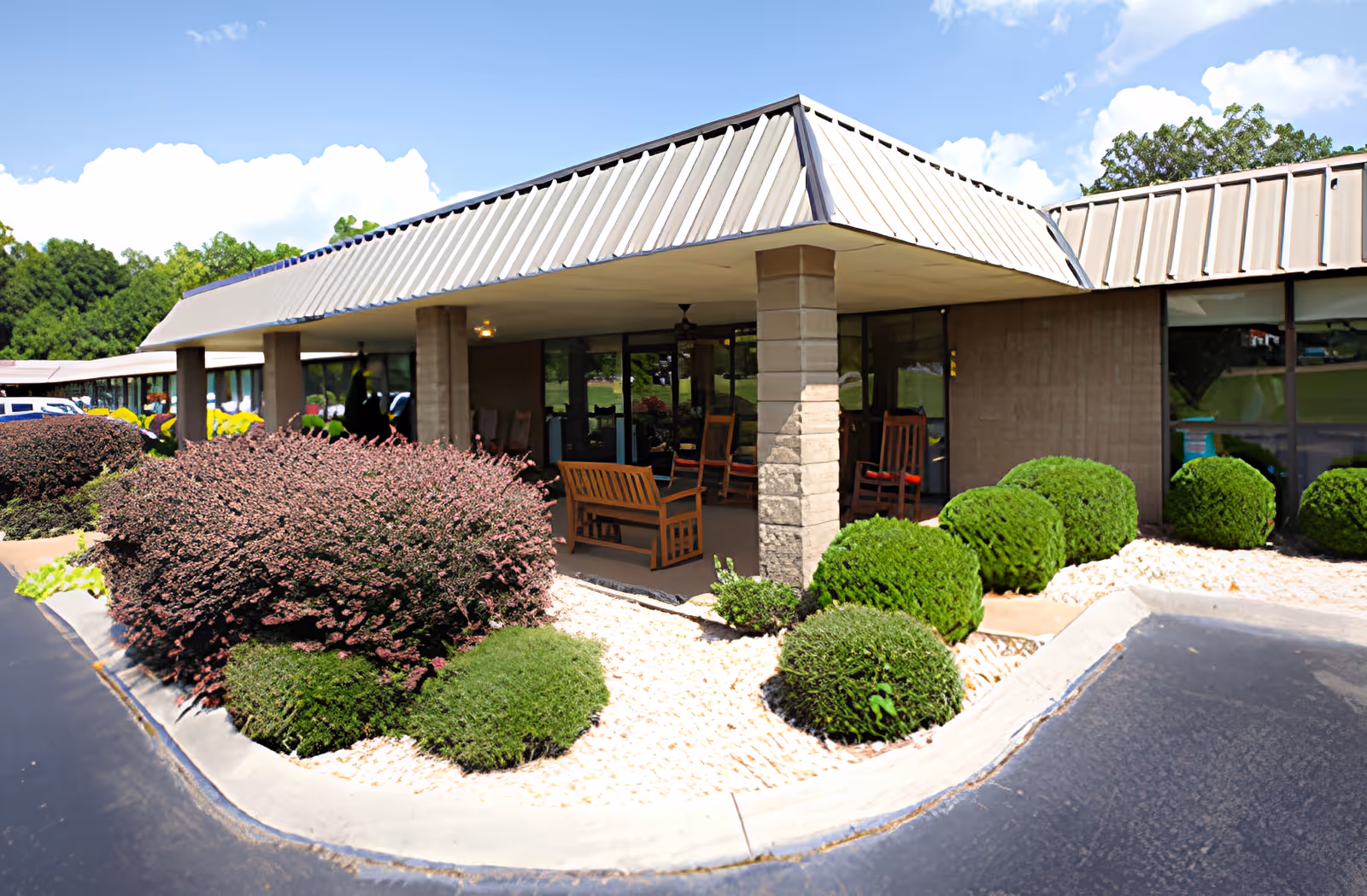 Exterior view of a single-story building with a covered porch area featuring wooden rocking chairs and benches. The building has large windows and a metal roof. The surrounding landscaping includes neatly trimmed bushes and white gravel beds under a bright blue sky with some clouds.