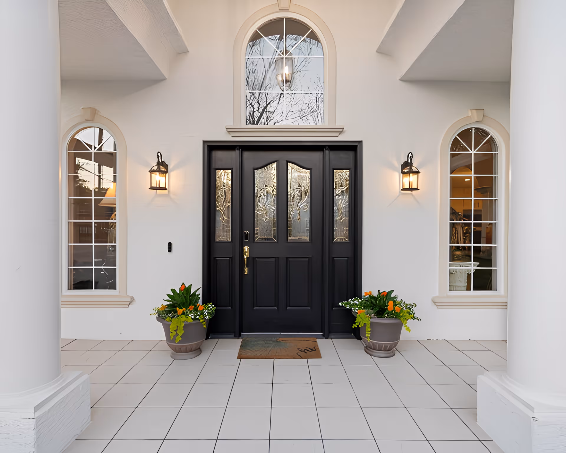 Front entrance of a building with a black double door featuring decorative glass panels, flanked by two tall arched windows. Two wall-mounted lantern-style lights are on either side of the door, and two large flower pots with green plants and orange flowers are placed symmetrically on the tiled floor. The exterior walls are white, and there are two large white columns visible on either side of the entrance.