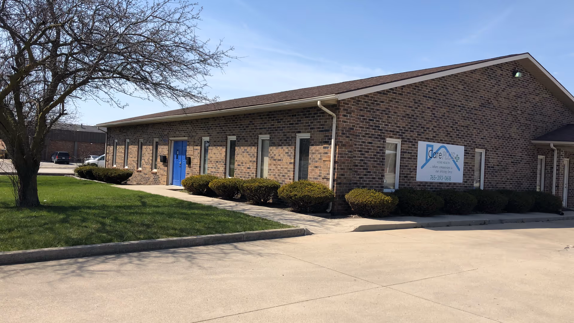 Exterior of a single-story brick facility with a blue door, sign, shrubs, and a parking area.