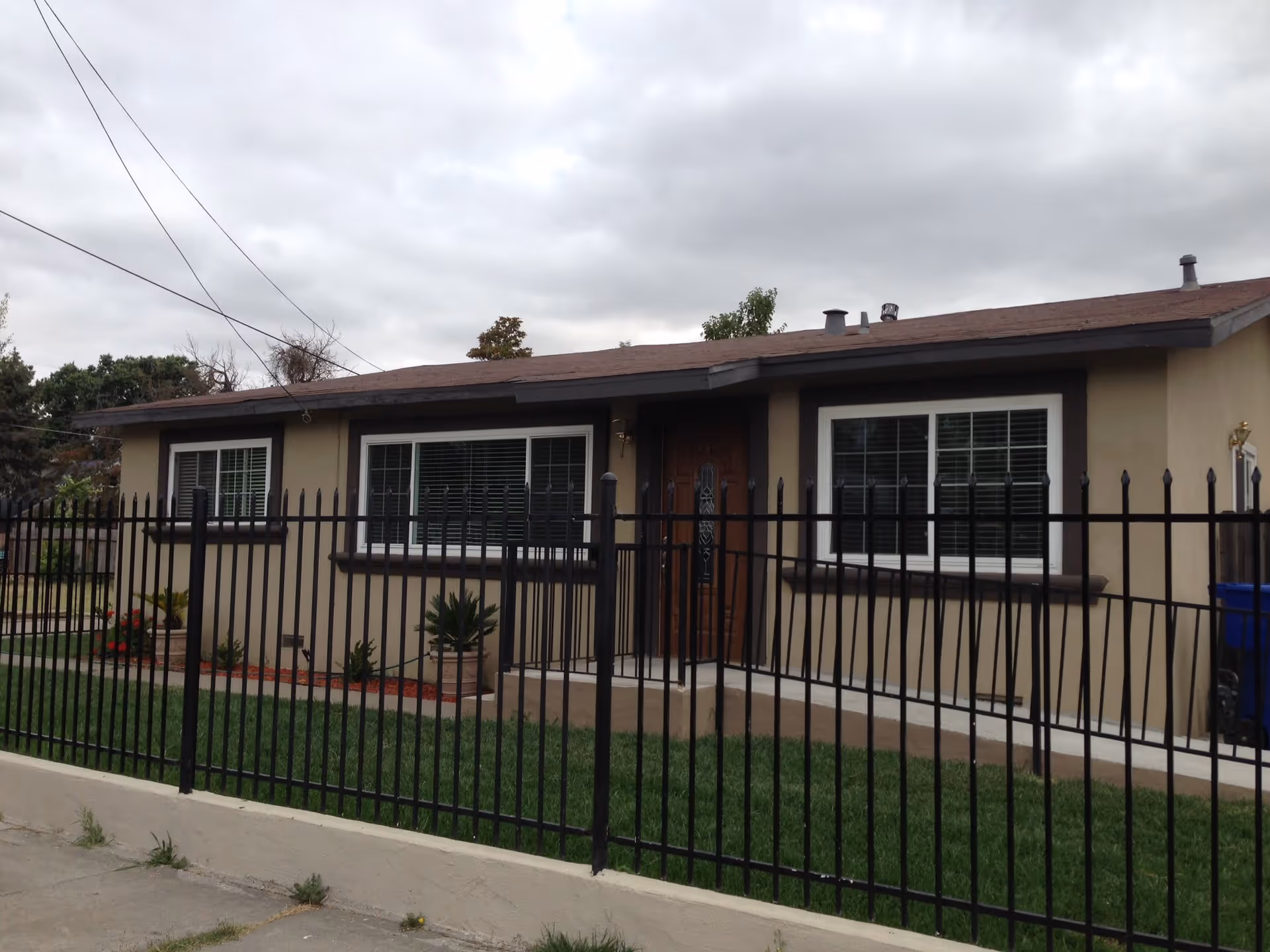 Single-story beige house with white-trim windows and a black metal fence in front under a cloudy sky.