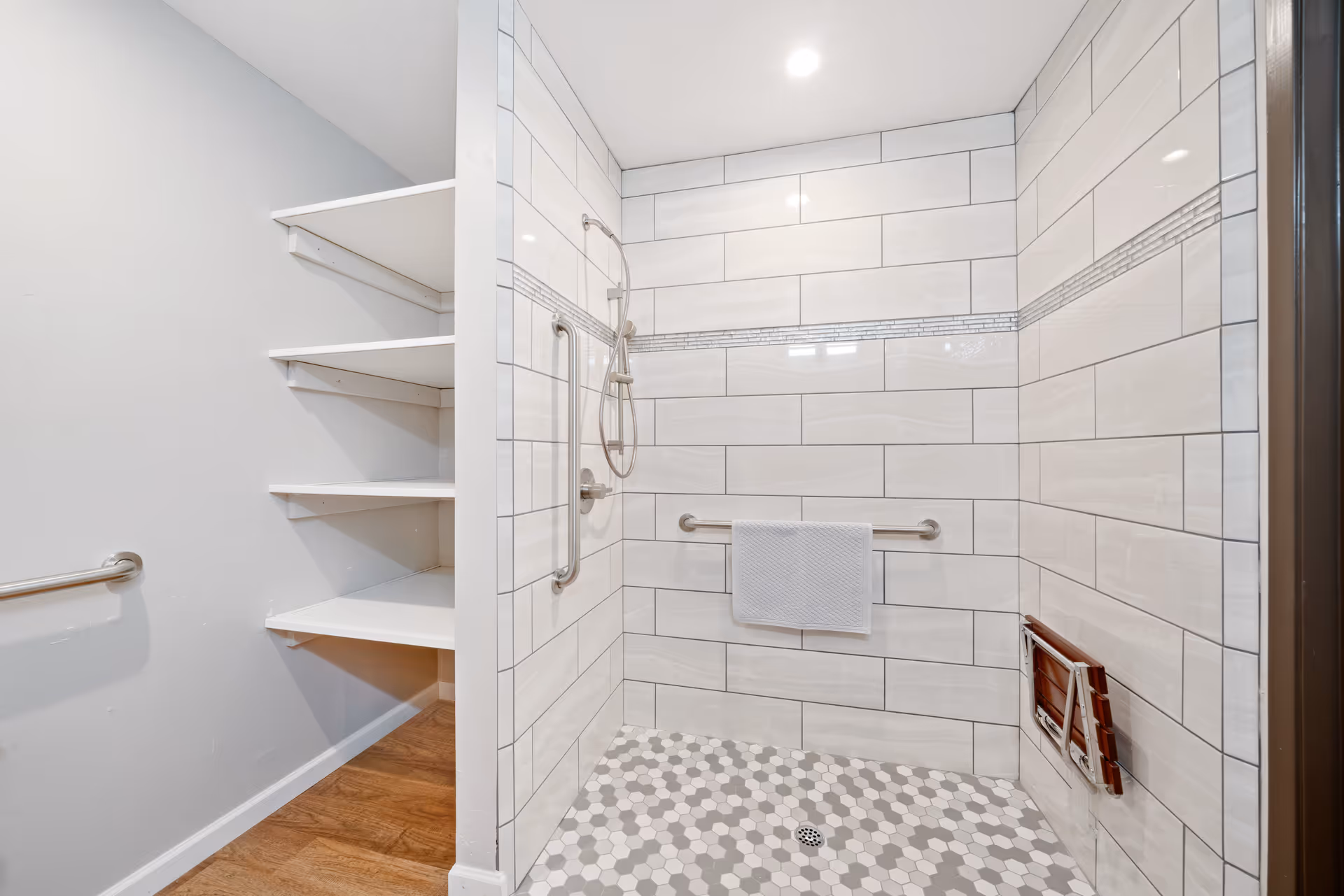 A modern walk-in shower with white and gray hexagonal floor tiles and large rectangular white wall tiles with a decorative horizontal strip. The shower features a handheld showerhead, two stainless steel grab bars, a towel hanging on one grab bar, and a foldable wooden shower seat mounted on the wall. Adjacent to the shower is an open shelving unit with white shelves and a wooden floor.