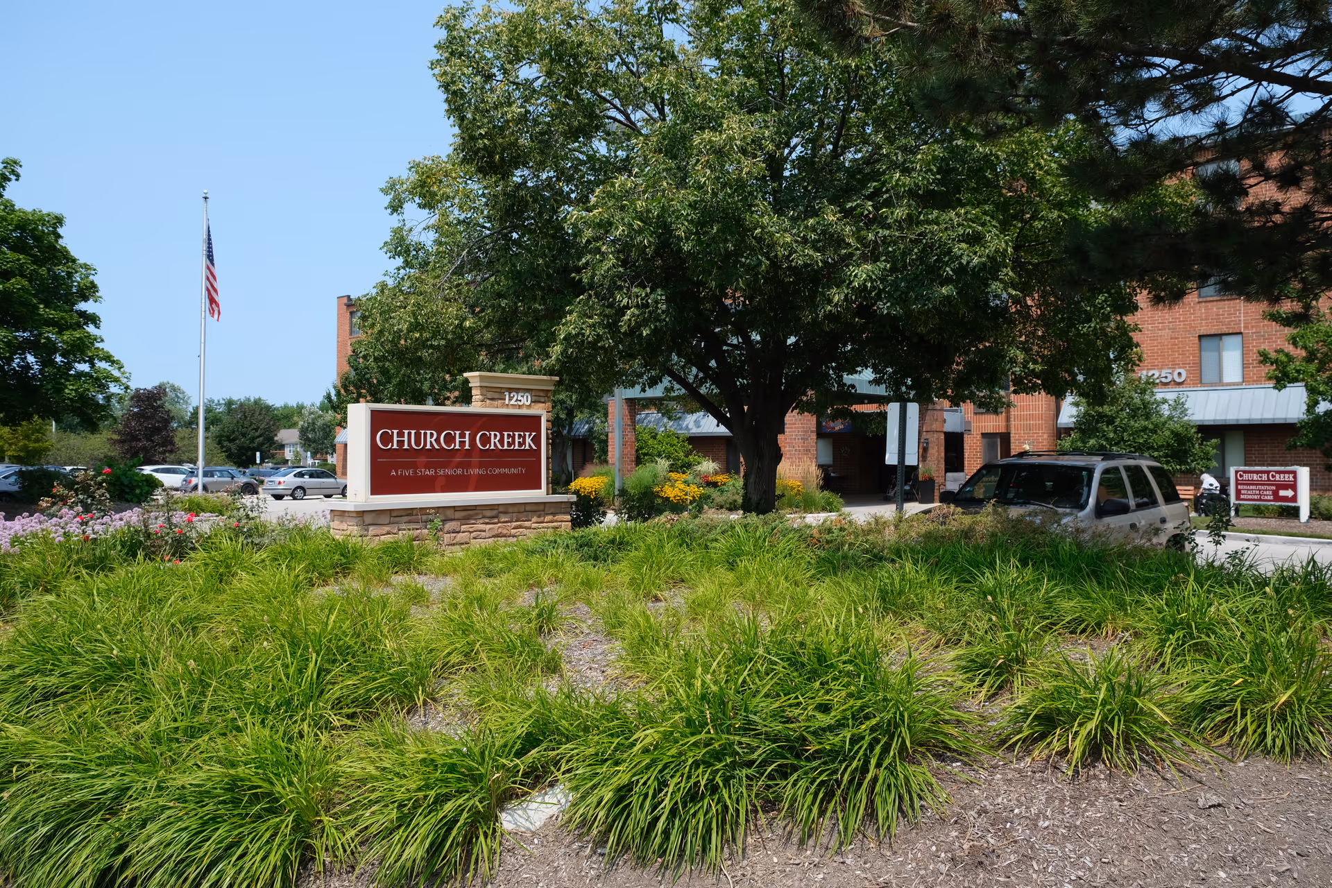 Front entrance of the Church Creek senior living community with a prominent sign, landscaped grounds, a large tree, and the building facade in the background.