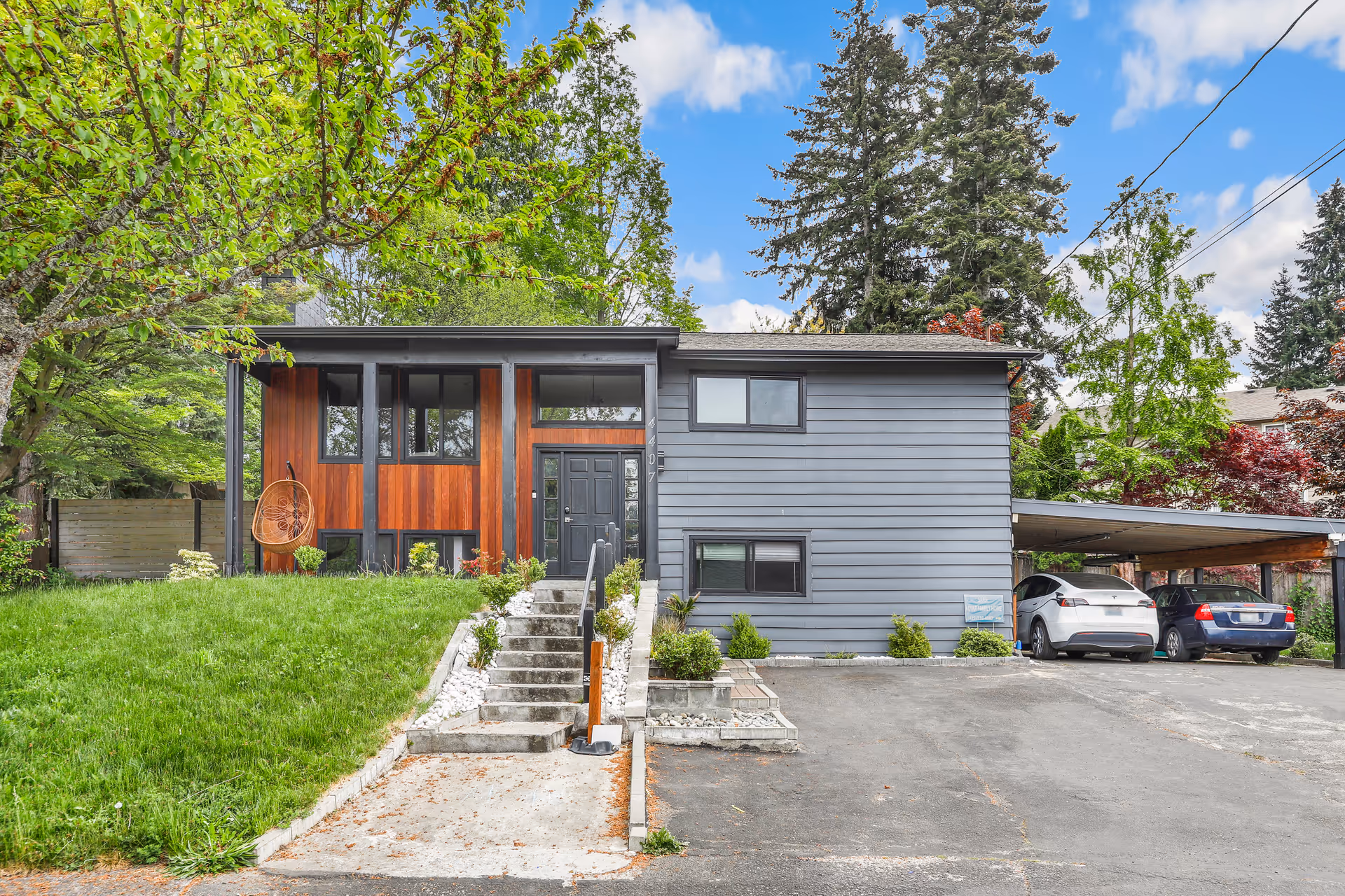 Modern, two-tone house with front entrance, steps, and carport amid green trees on a sunny day.