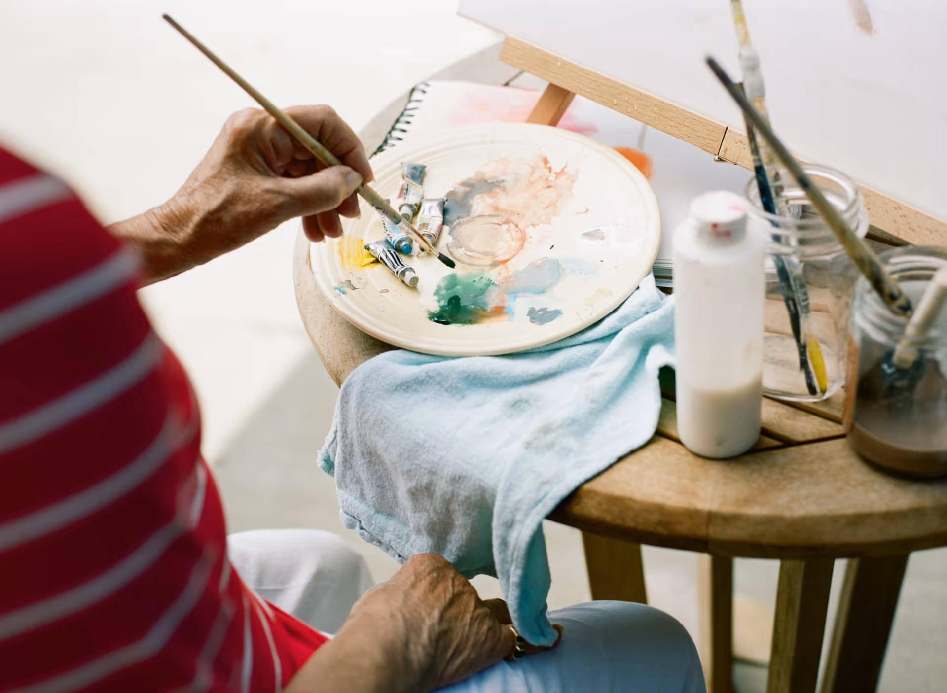 A person paints at a table with a paint palette, brushes, and jars.