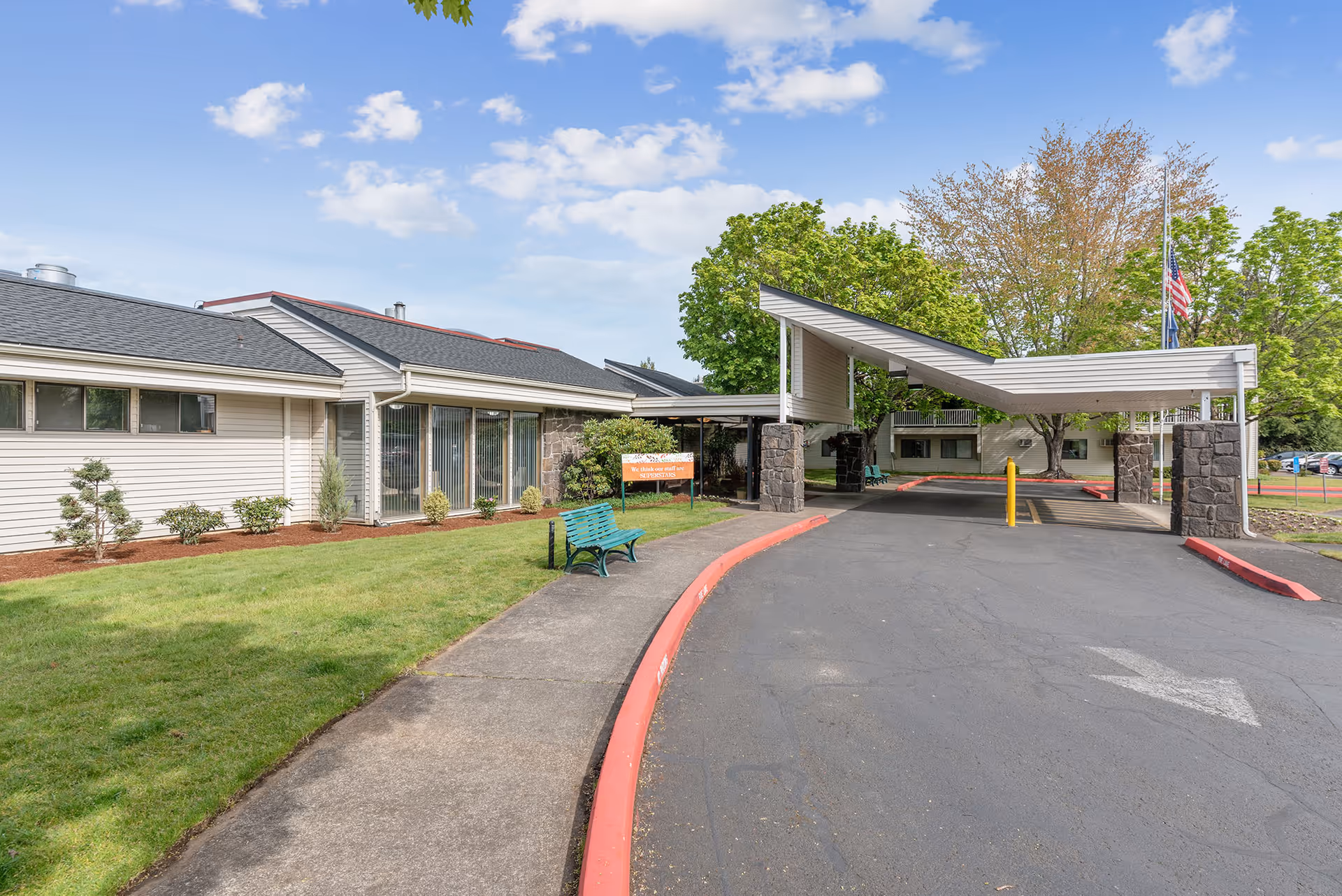 Front driveway and covered porte-cochère of a senior living building with a green bench, lawn, and an American flag.