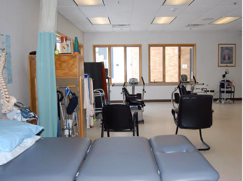 Rehabilitation/exercise therapy room with treatment tables, exercise bikes, chairs, walkers and medical equipment.