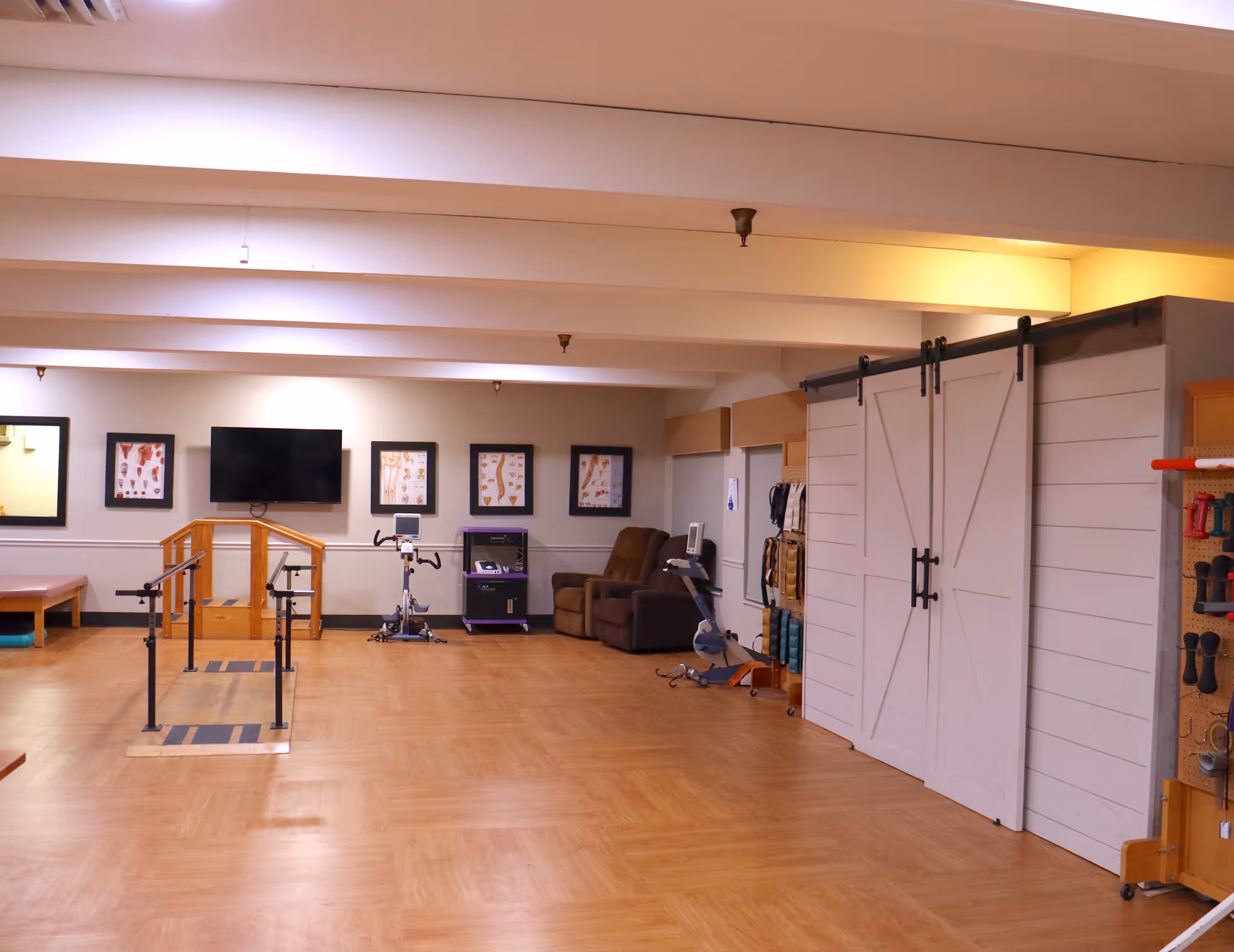 Spacious rehabilitation/activity room with parallel walking bars, exercise equipment, a TV, seating, and sliding barn doors.