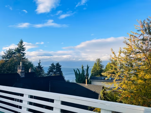 A seaside view from a balcony with white railing, rooftops, trees, and the ocean.