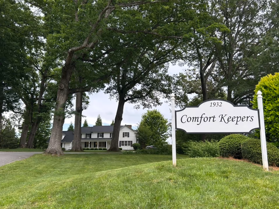 A large Comfort Keepers sign in the foreground with a white house and tall trees behind.