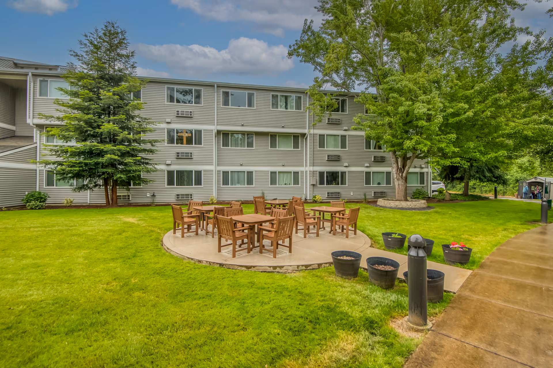 Outdoor courtyard with a circular seating area of wooden tables and chairs on a lawn in front of a three-story apartment building and trees.