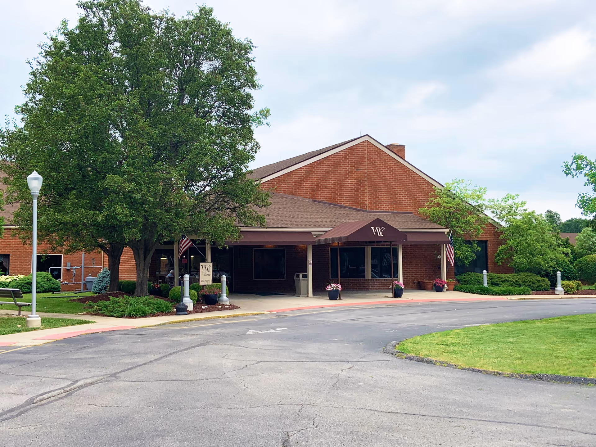 Exterior view of Westminster Village Kentuckiana, a single-story brick building with a covered entrance. There are trees, shrubs, and a well-maintained lawn surrounding the building. Two American flags are displayed near the entrance, and a sign with the facility's name is visible near the walkway.