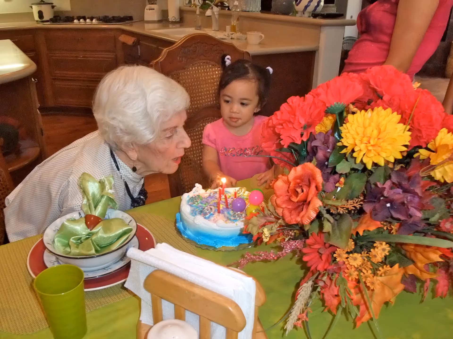 An elderly woman and a young girl are sitting at a dining table in a kitchen. The elderly woman is blowing out candles on a birthday cake while the girl watches. The table is decorated with a large colorful bouquet of flowers and tableware.