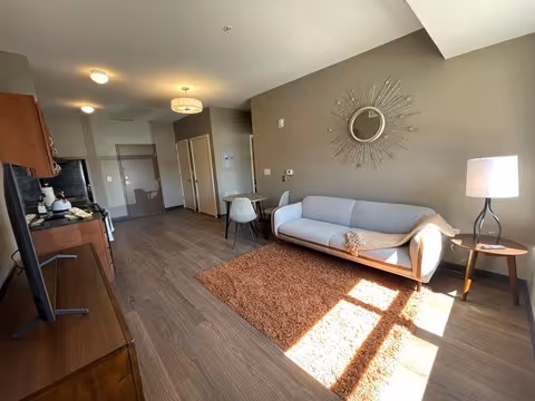 Sunlit studio living area with a sofa under a decorative round mirror, a rug, side table with lamp, small dining table and kitchenette along one wall.