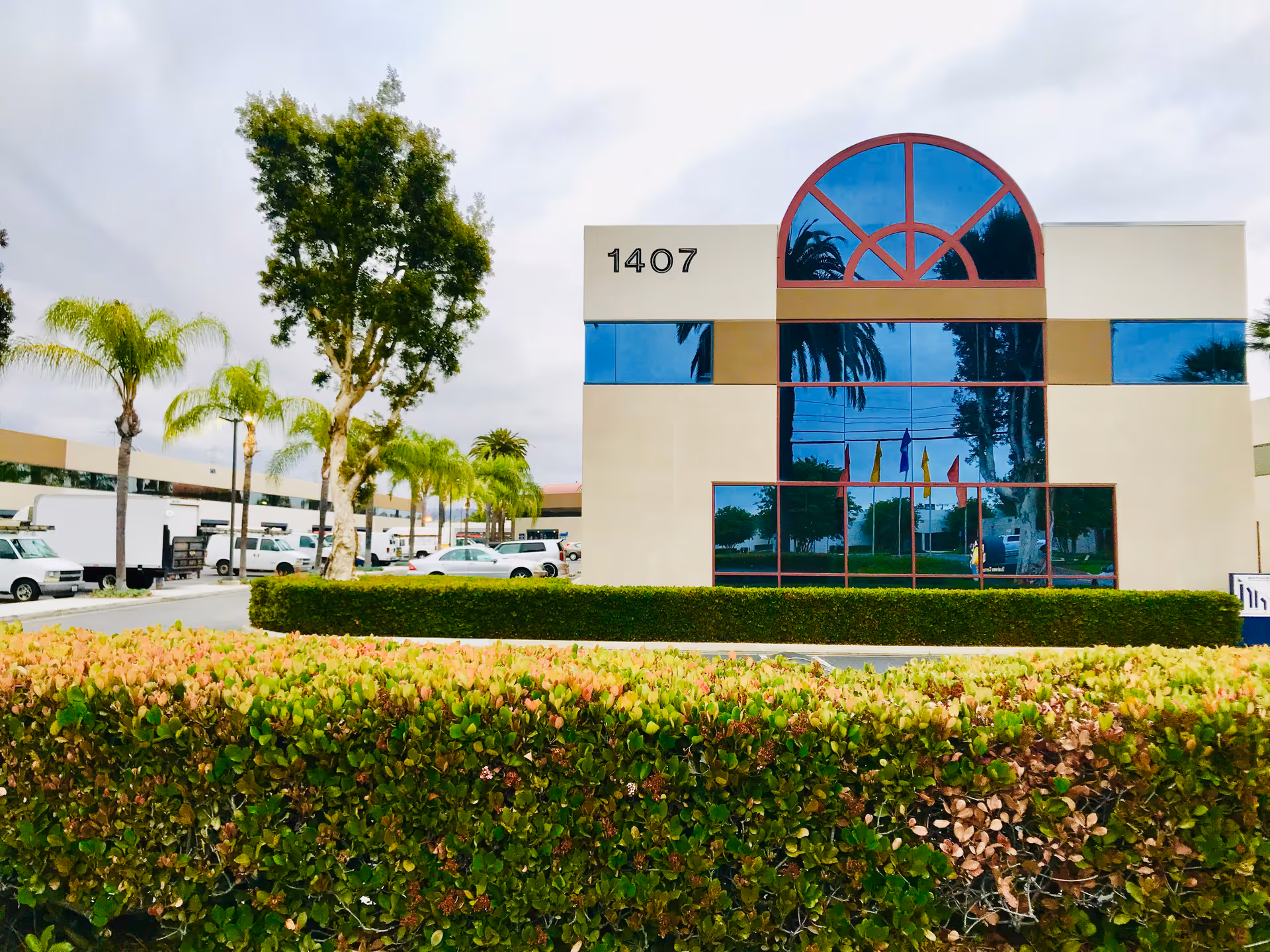Front view of a modern facility with large glass windows, palm trees, and trimmed hedges.
