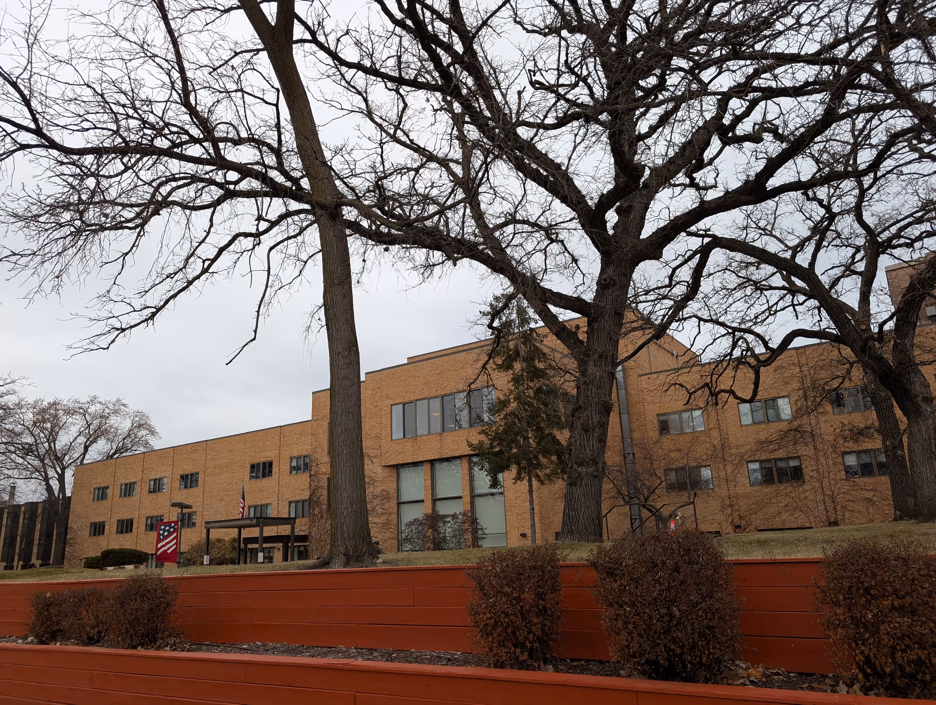 Exterior view of a large brick building with multiple windows, leafless trees in the foreground, and a tiered red retaining wall with bushes. The sky is overcast and an American flag is visible near the entrance.