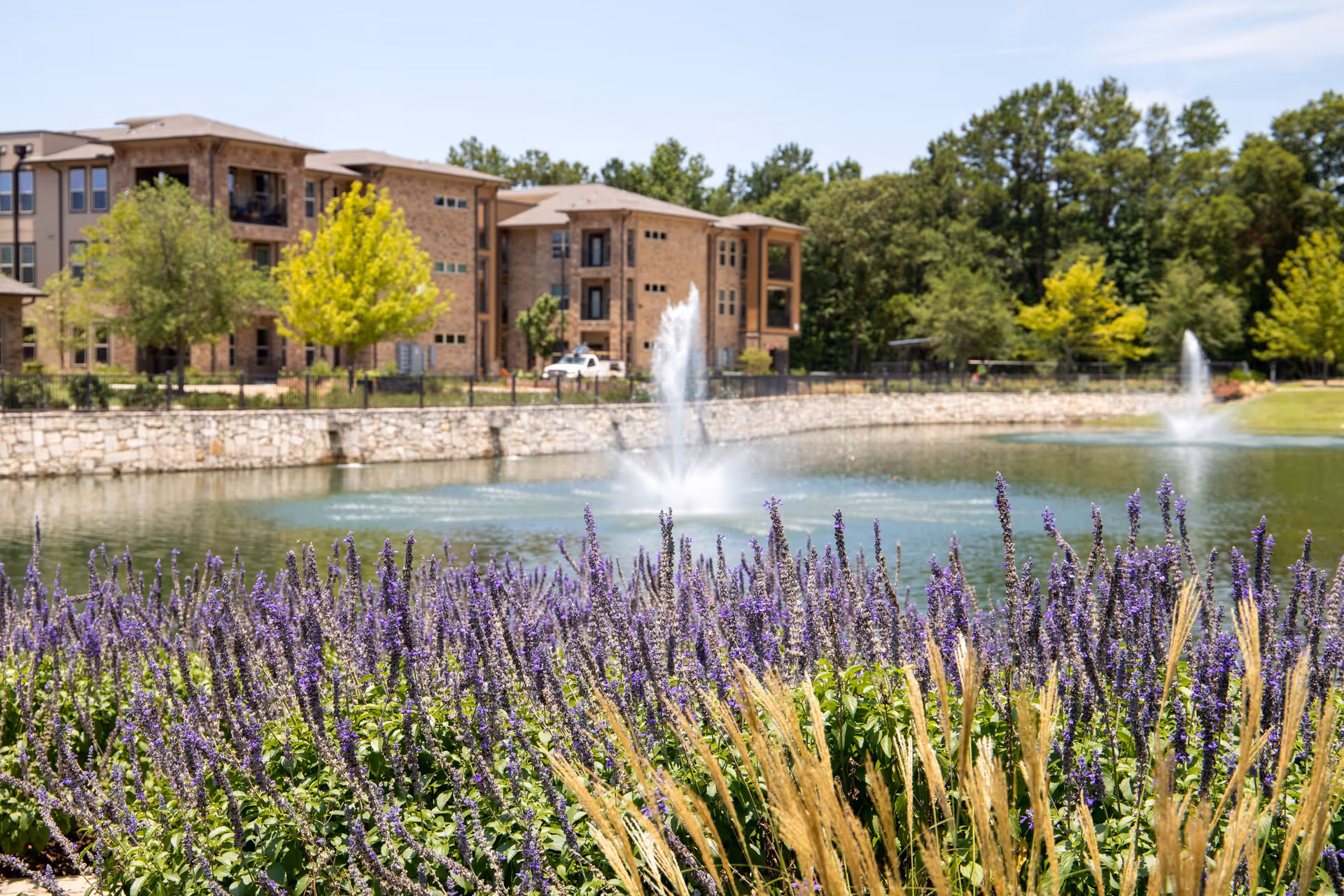 Purple flowers and ornamental grasses by a pond with fountains in front of a multi-story brick residential building and trees.