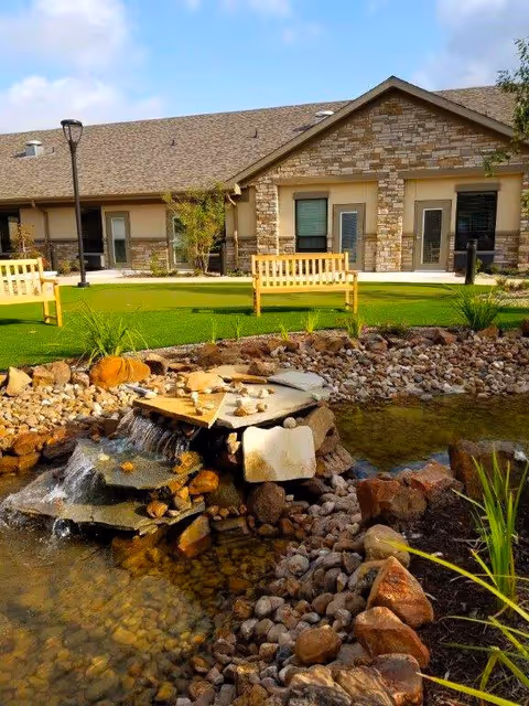 Outdoor garden area at Cedar Pointe Wellness And Health Center featuring a small artificial waterfall flowing over rocks into a pond, surrounded by landscaping rocks and plants. In the background, there are two wooden benches on a green lawn with a stone and brick building behind them under a partly cloudy sky.