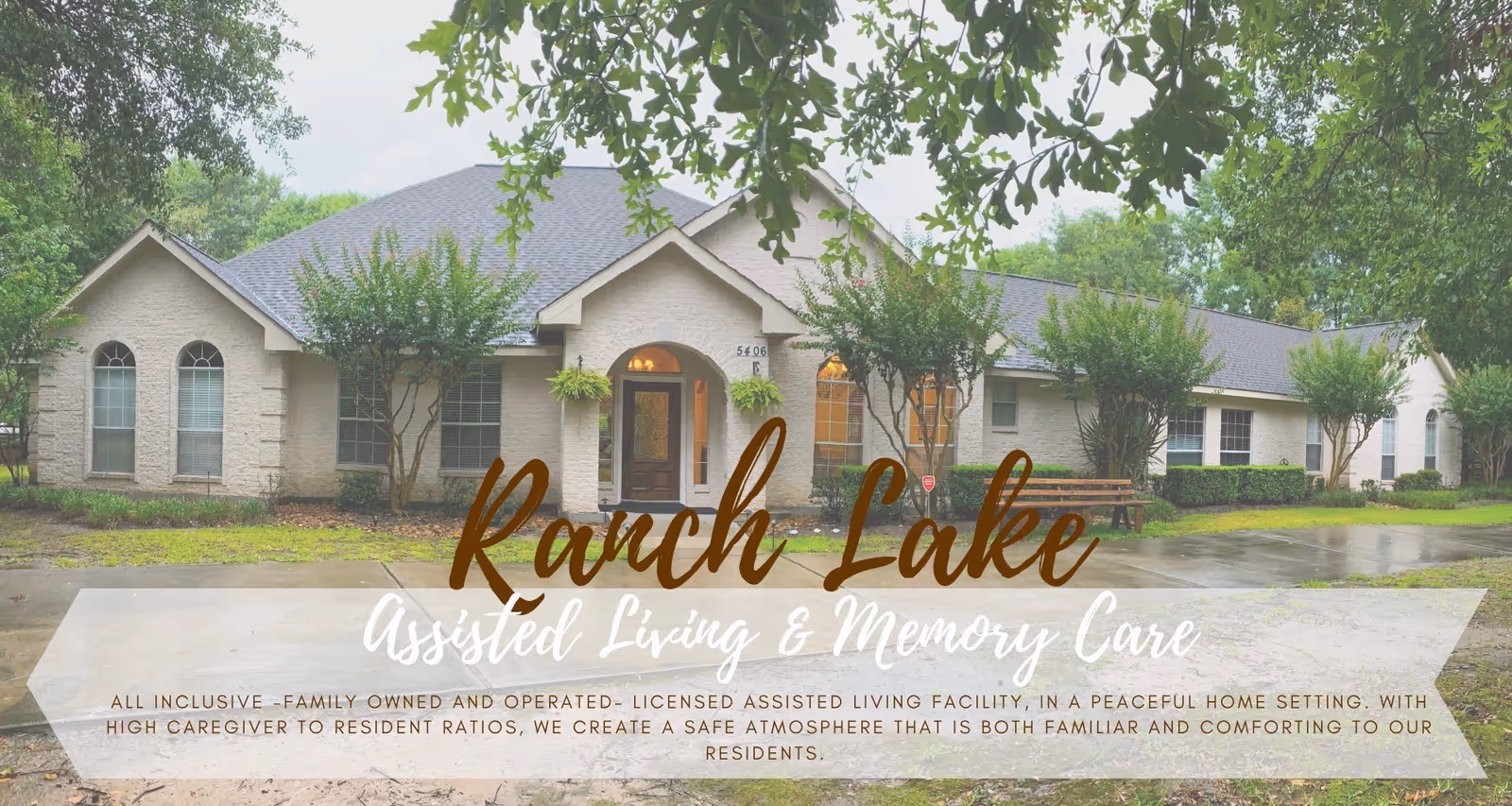 Exterior view of Ranch Lake Assisted Living facility showing a single-story building with a gray roof, beige brick walls, several windows, and a front entrance with a small porch. The building is surrounded by trees and greenery, with a bench and a paved walkway in front.