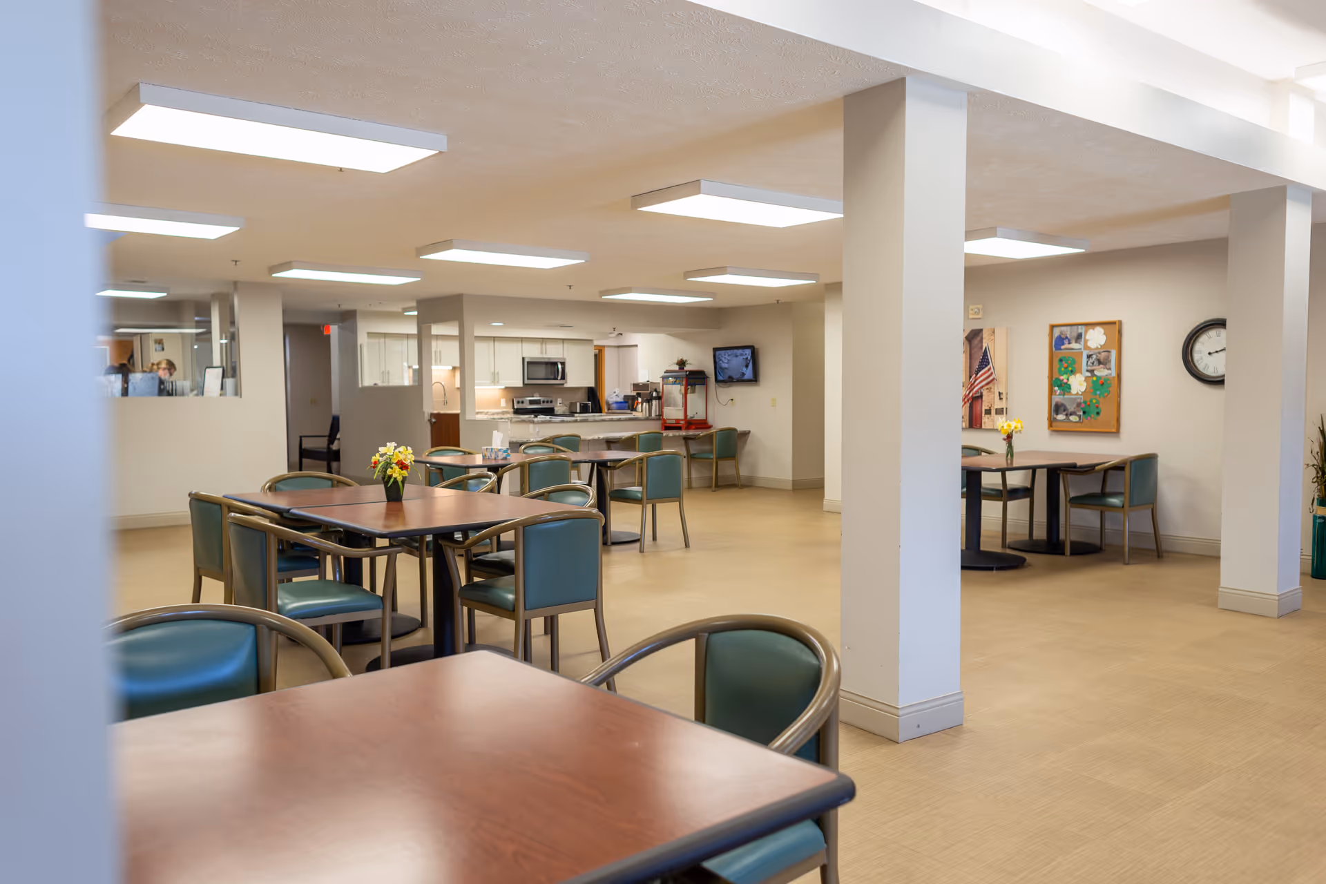 Bright communal dining area with tables and chairs; kitchen visible in the background.