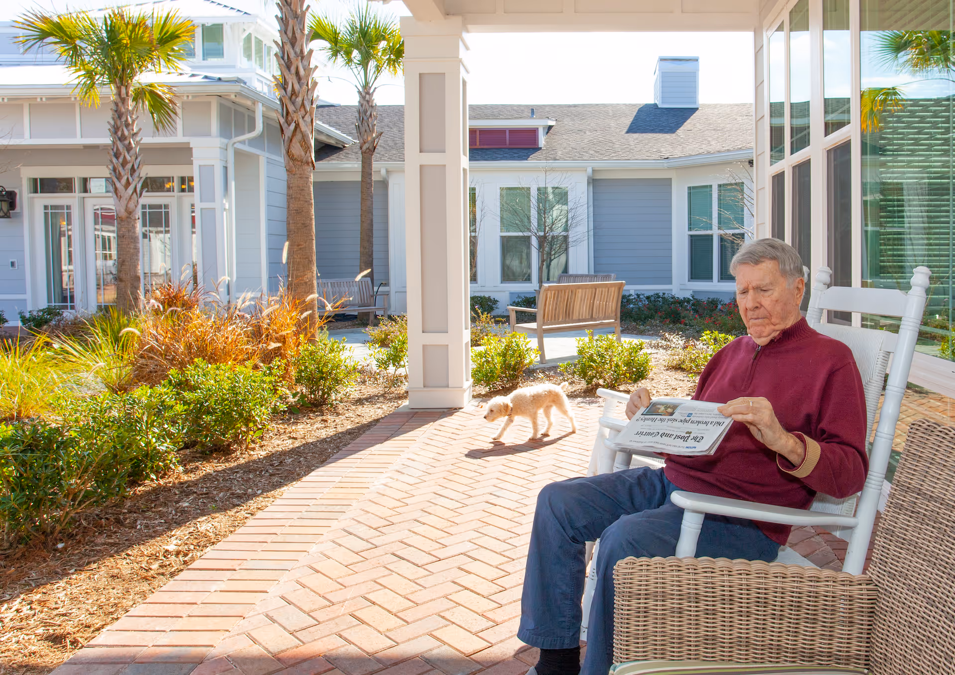 An elderly man wearing a maroon sweater and blue pants sits on a white rocking chair on a covered patio, reading a newspaper. A small dog walks nearby on the brick-paved patio. The background shows a well-maintained garden with bushes, palm trees, and a bench, with light gray buildings and large windows under a clear sky.