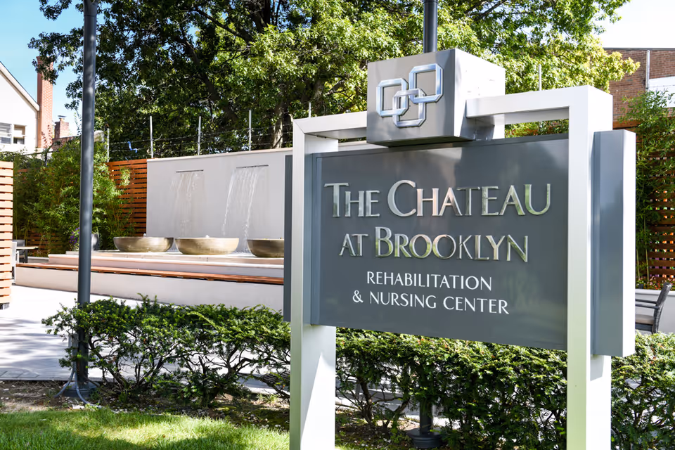 Outdoor view of a sign for The Chateau at Brooklyn Rehabilitation & Nursing Center with a decorative water feature and greenery in the background.