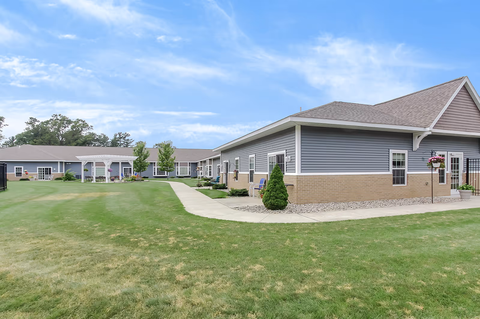 Exterior view of a single-story assisted living facility with manicured lawn, sidewalks, and a covered pavilion.