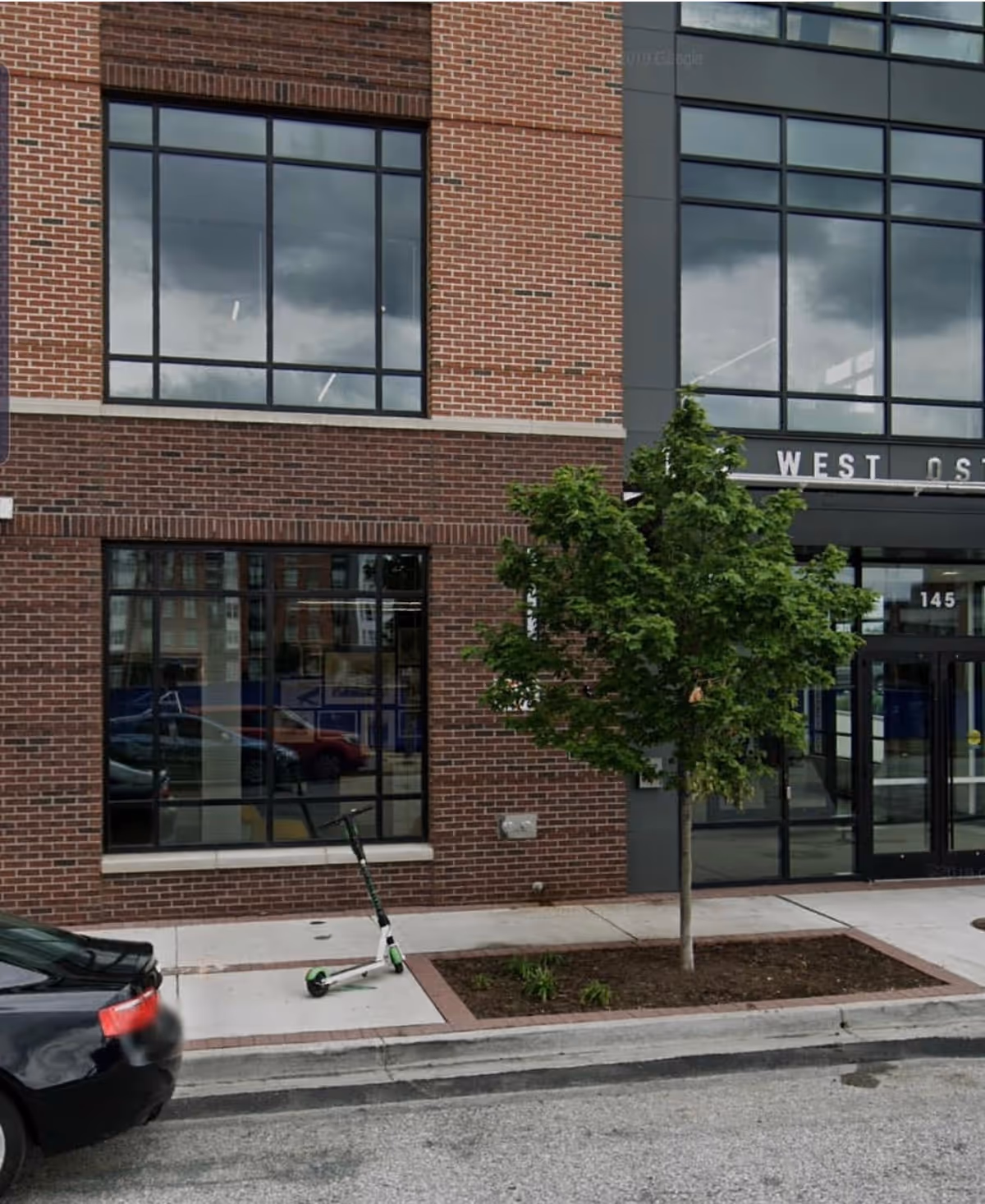 Brick building exterior with large storefront windows, a tree, and a parked scooter by the sidewalk.