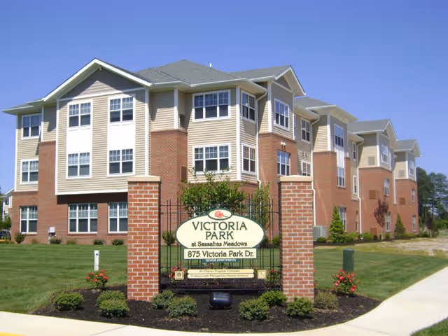 Exterior front of a multi-story brick-and-siding senior living building with landscaping and a sign reading 'Victoria Park at Sassafras Meadows'.