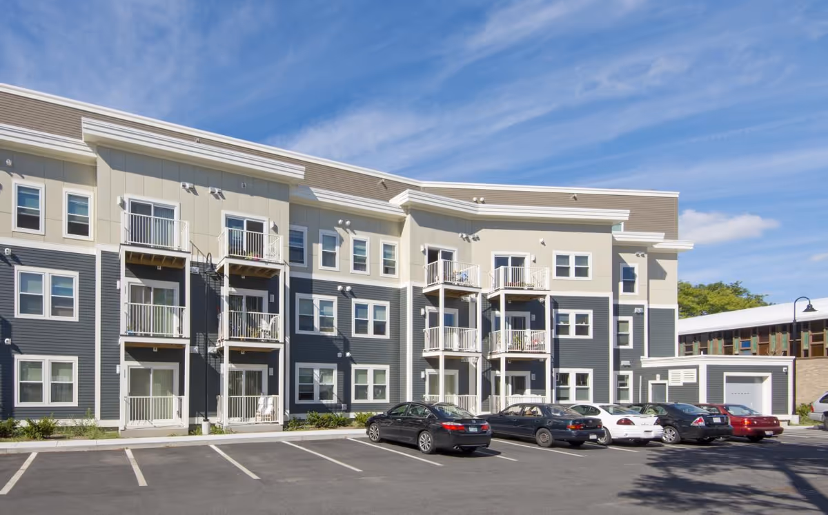 Modern multi-unit senior living building with balconies and a parking lot under a blue sky.