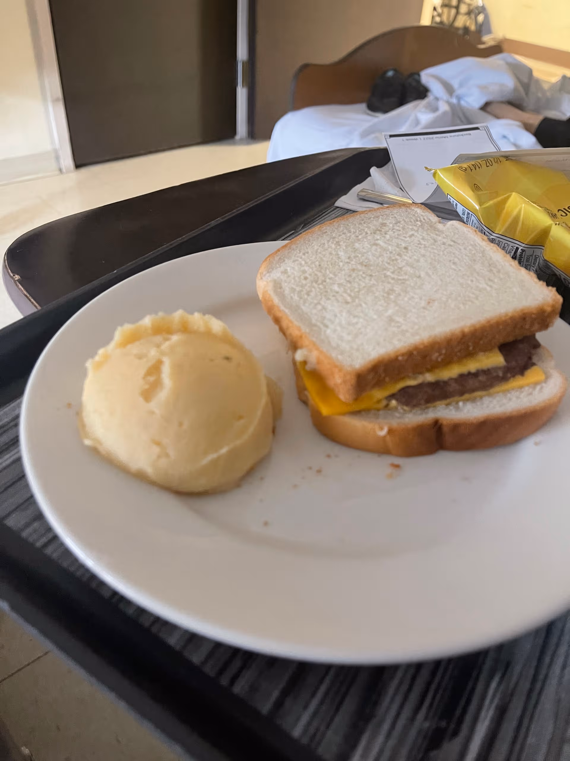 Plate with sandwich and mashed potatoes on a tray; a hospital bed behind in the background.