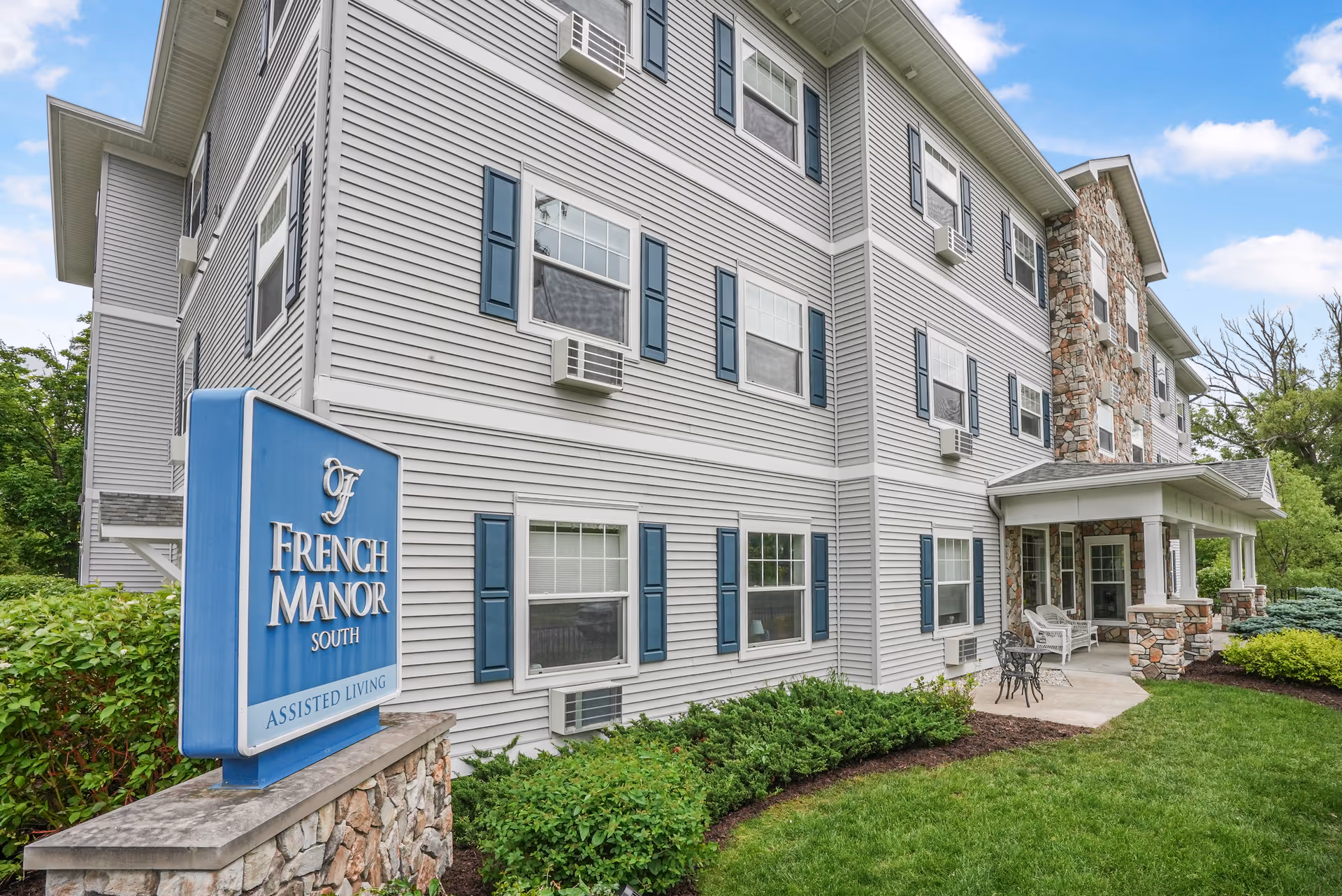 Three-story gray siding senior living building with blue shutters and a front entrance sign.