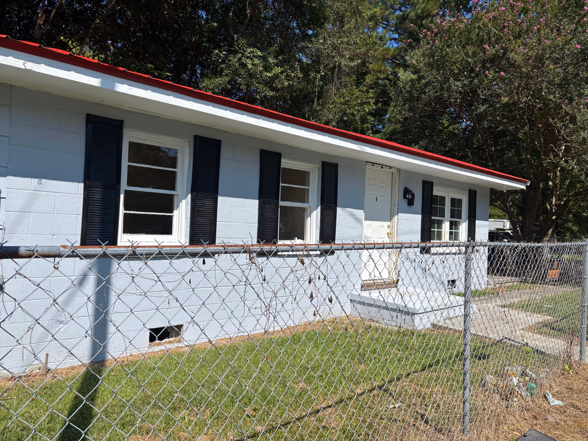 Single-story light-blue building with black shutters, red trim, and a chain-link fence.