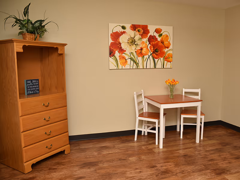 Small dining nook with a two-seat wooden table and chairs beside a wooden dresser under a floral painting.