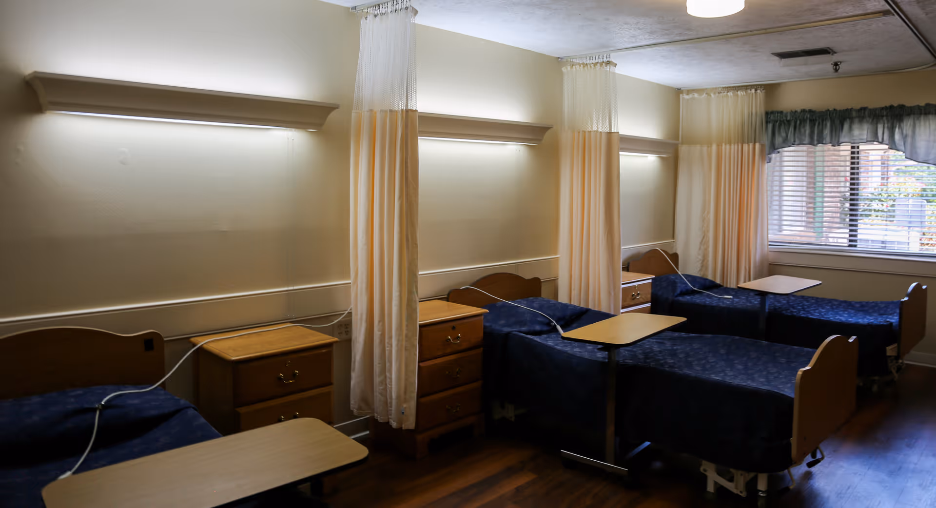 A room with three hospital-style beds separated by privacy curtains. Each bed has a wooden nightstand and an overbed table. The beds have dark blue bedding, and there is a window with blinds and a valance on the right side of the room. The floor is wooden, and the walls are light-colored with overhead lighting above each bed.