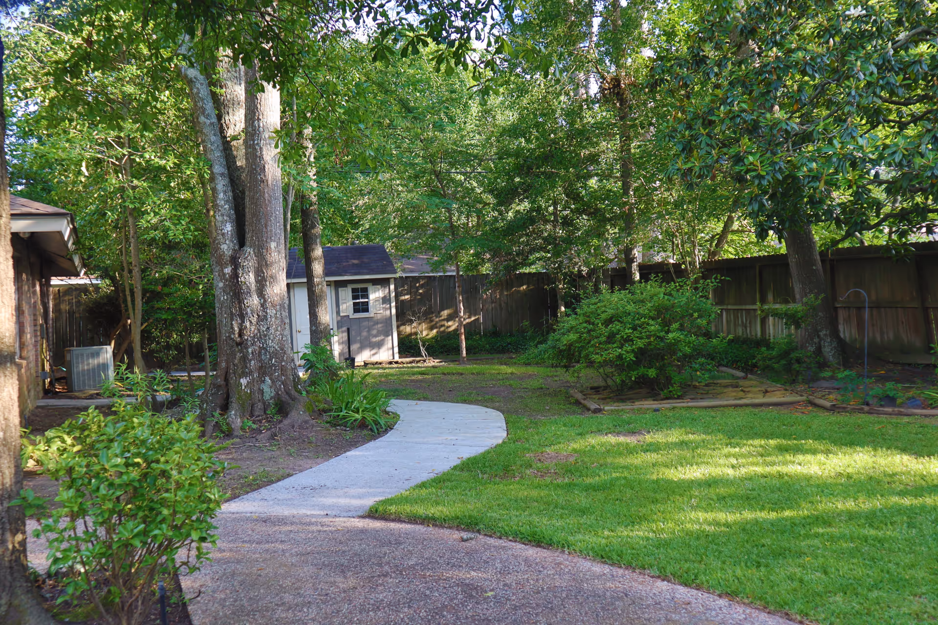 A backyard with a curved concrete path leading to a small shed, surrounded by trees, lawn, and a wooden fence.