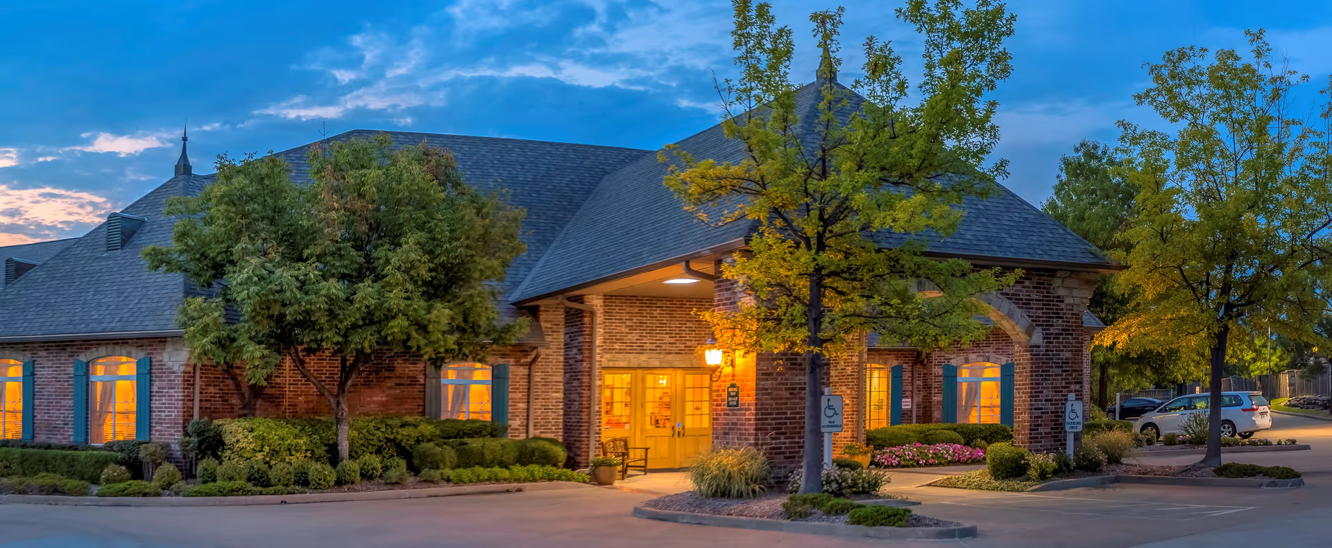 Exterior of The Parke Assisted Living building at dusk with lit entrance and trees.