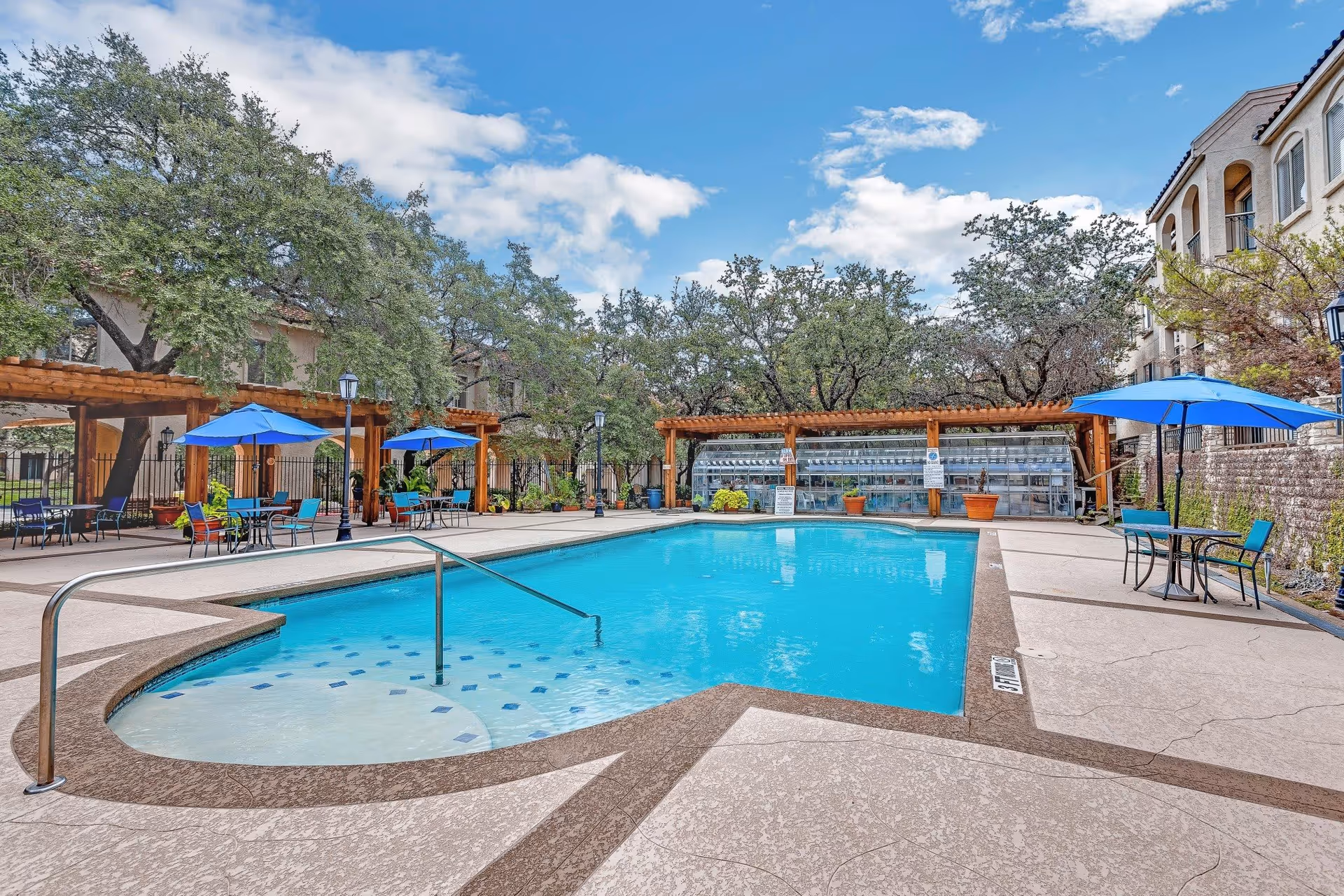 Outdoor swimming pool area at Madison Estates with clear blue water, surrounded by lounge chairs, tables with blue umbrellas, pergolas, and trees under a partly cloudy sky.