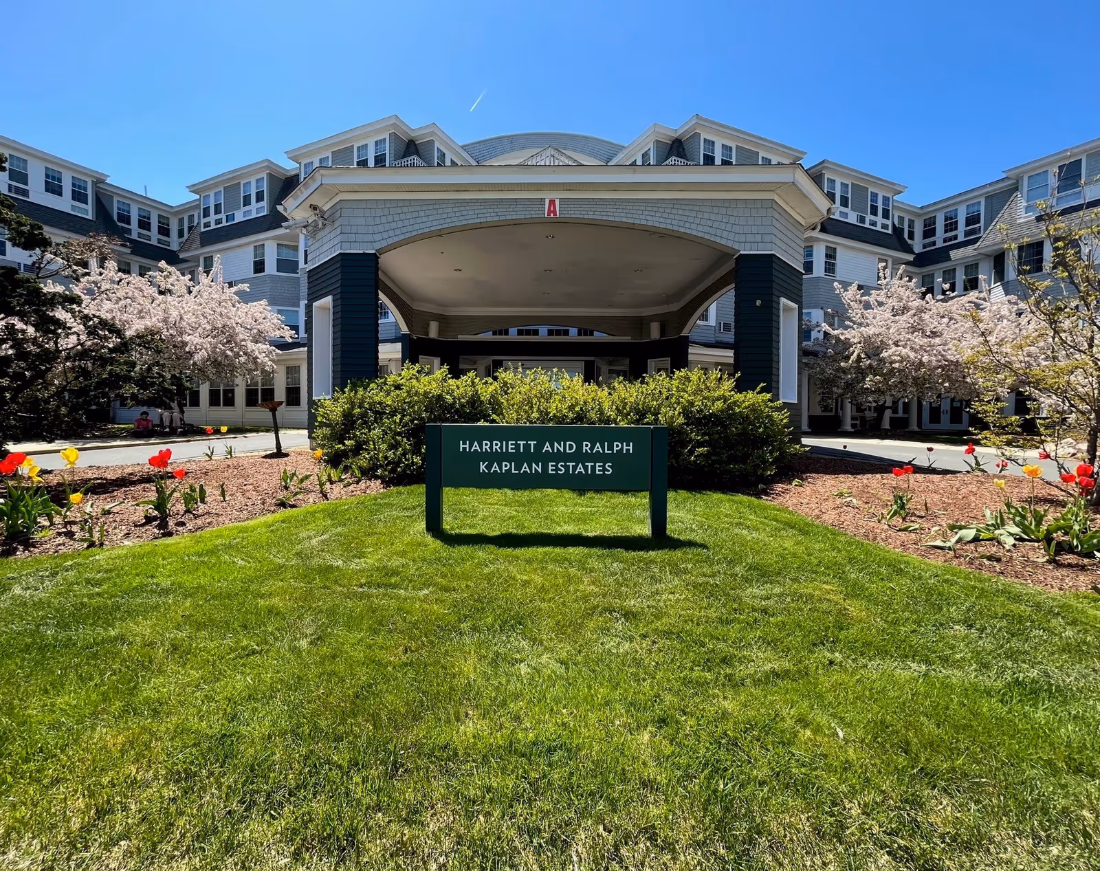 Front entrance of the Harriett and Ralph Kaplan Estates building with a covered drive, green sign, lawn and spring flower beds.