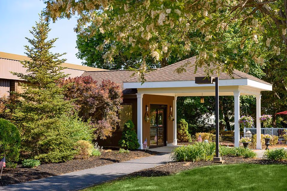 Entrance of a senior living facility with a covered porch supported by white columns, surrounded by well-maintained landscaping including green grass, shrubs, and trees. Small American flags are placed along the walkway leading to the glass door entrance.