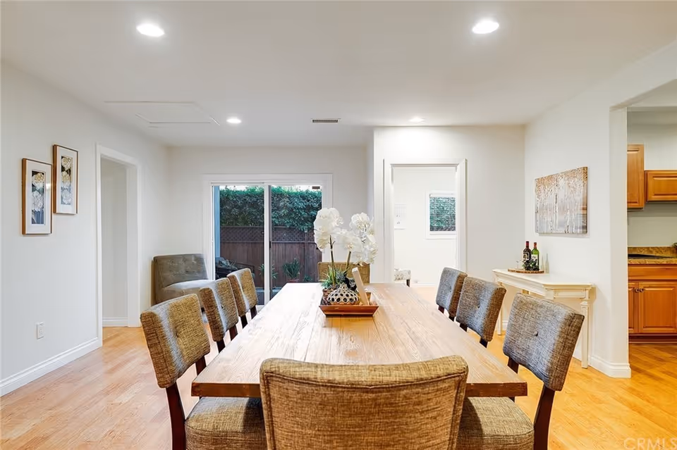 Bright dining room with a long wooden table surrounded by upholstered chairs and a sliding glass door to a fenced backyard.