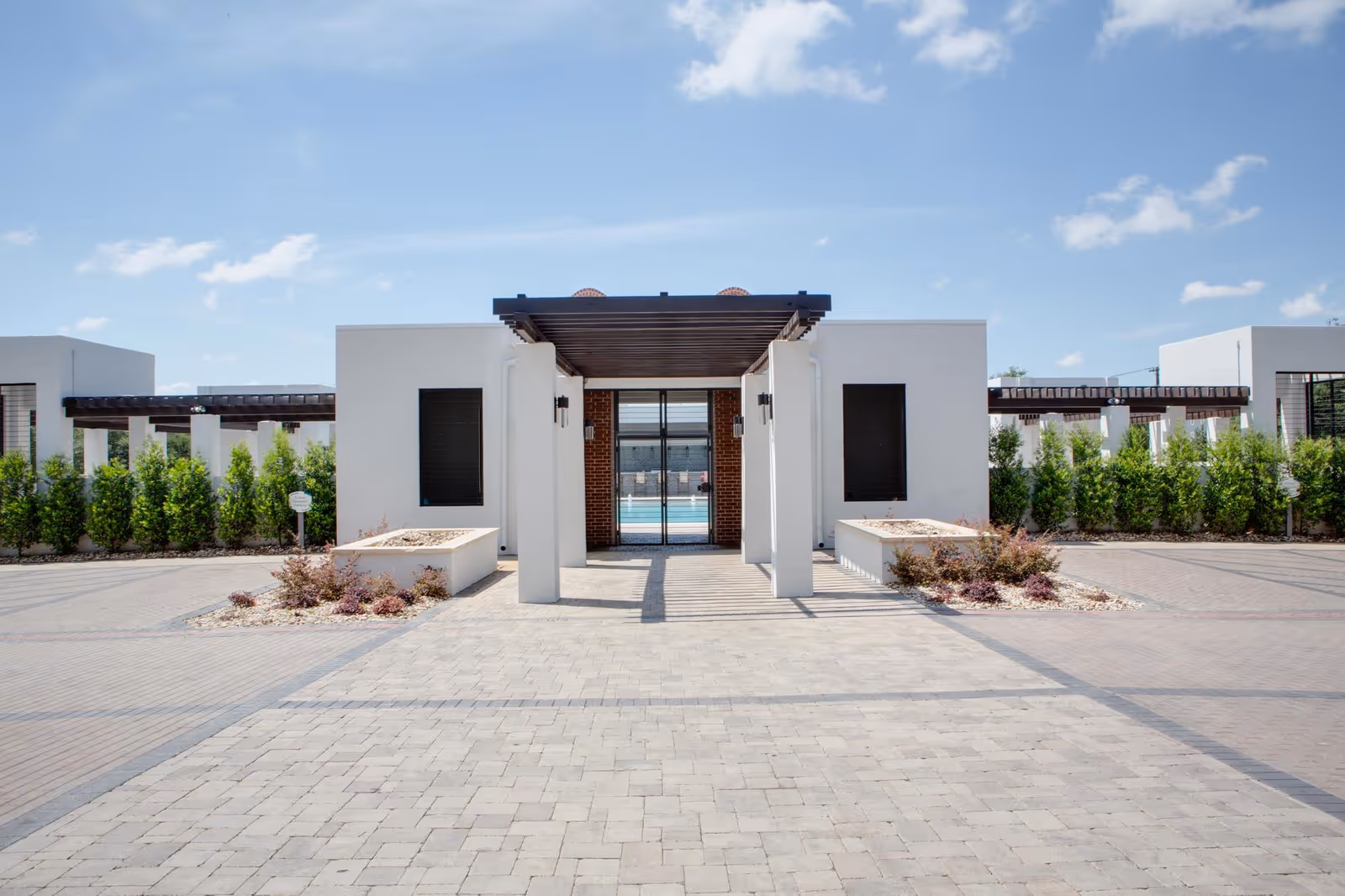 Modern white building entrance with a walkway, pergola, and neatly trimmed shrubs along the sides.