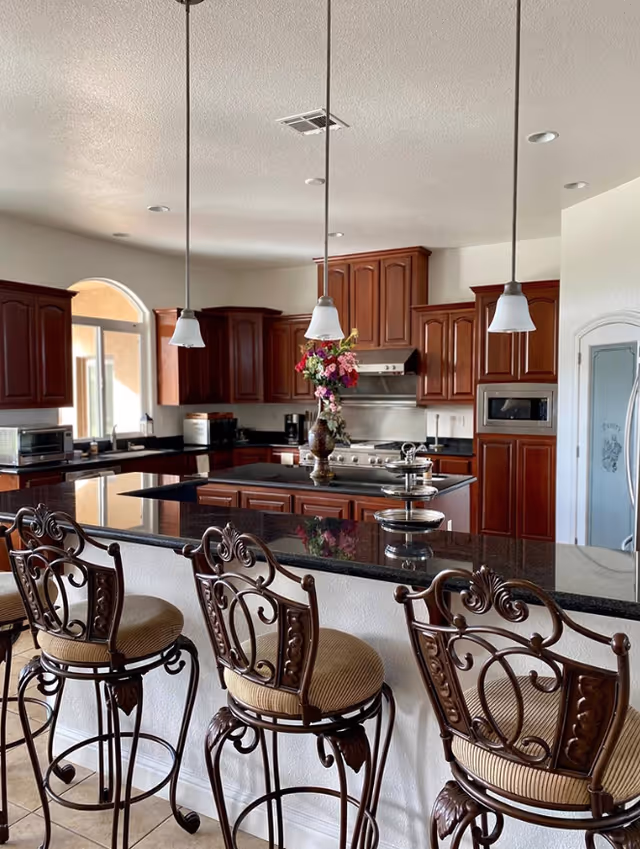 A spacious kitchen with dark granite countertops and wooden cabinets. Three ornate metal bar stools with cushioned seats are positioned at the kitchen island. Three pendant lights hang from the ceiling above the island. A vase with colorful flowers and a tiered tray are placed on the island. Stainless steel appliances including a stove, microwave, and toaster oven are visible. The kitchen has a bright window and a pantry door with frosted glass.