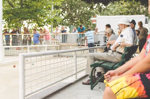 Elderly residents sit along an outdoor fenced court watching a game.