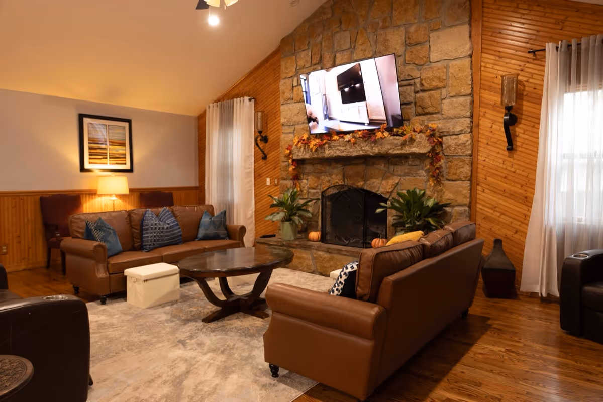 Cozy living room lounge with leather sofas, stone fireplace, mounted TV, and wooden paneling.