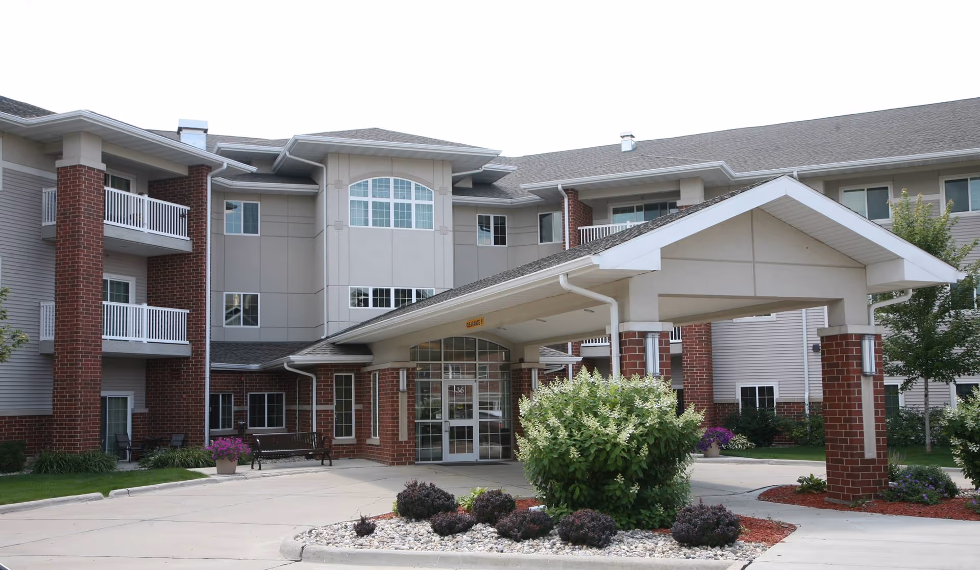 Front entrance of a multi-story senior living building with a covered porte-cochere, brick columns, balconies, and landscaped driveway.